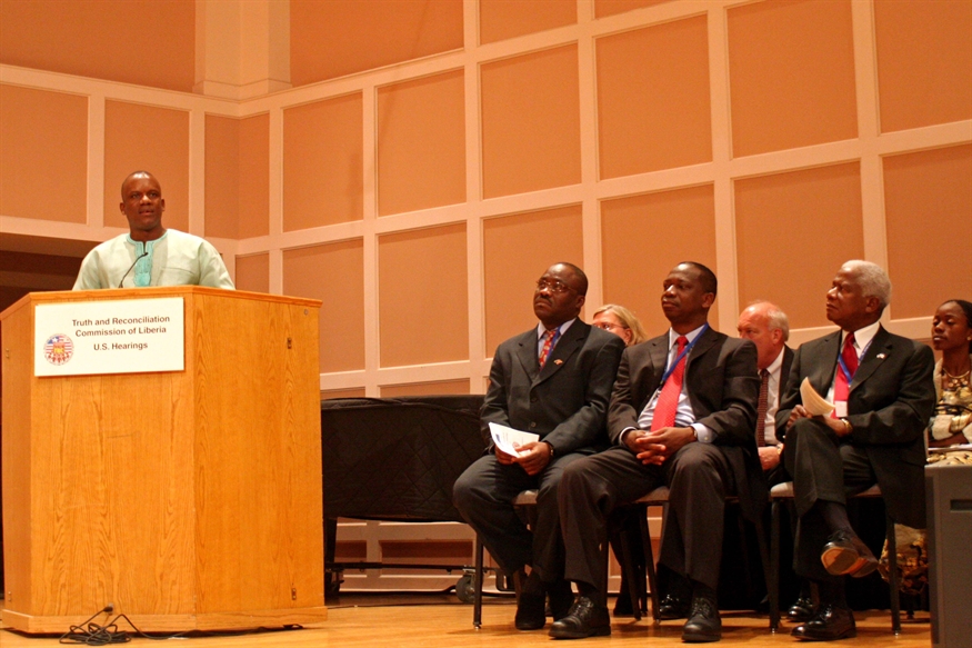 [ai] A speaker at a podium delivers a speech, while four seated men listen attentively. The setting appears to be a formal event, with a wooden stage and a banner reading 'Truth and Reconciliation Commission of Liberia'.